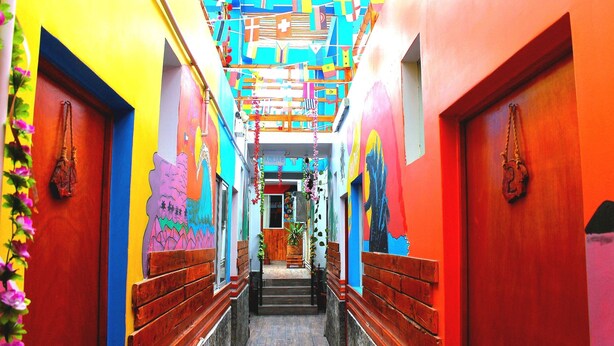 A narrow hallway with colorful walls and doors, leading to a central area with a wooden bench and potted plants. - Raymi House Hostel (Arica)