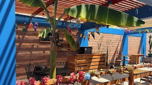 An outdoor patio with wooden tables and chairs, a brick wall, and a blue awning. - Raymi House Hostel (Arica)