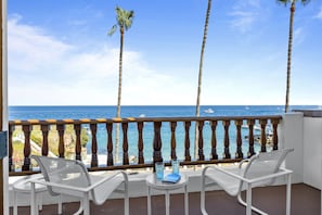 A balcony with two white chairs and a small table, overlooking a beach with palm trees and boats.