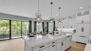 Een moderne keuken met witte kasten, een groot eiland, hanglampen en uitzicht op groen door grote ramen.