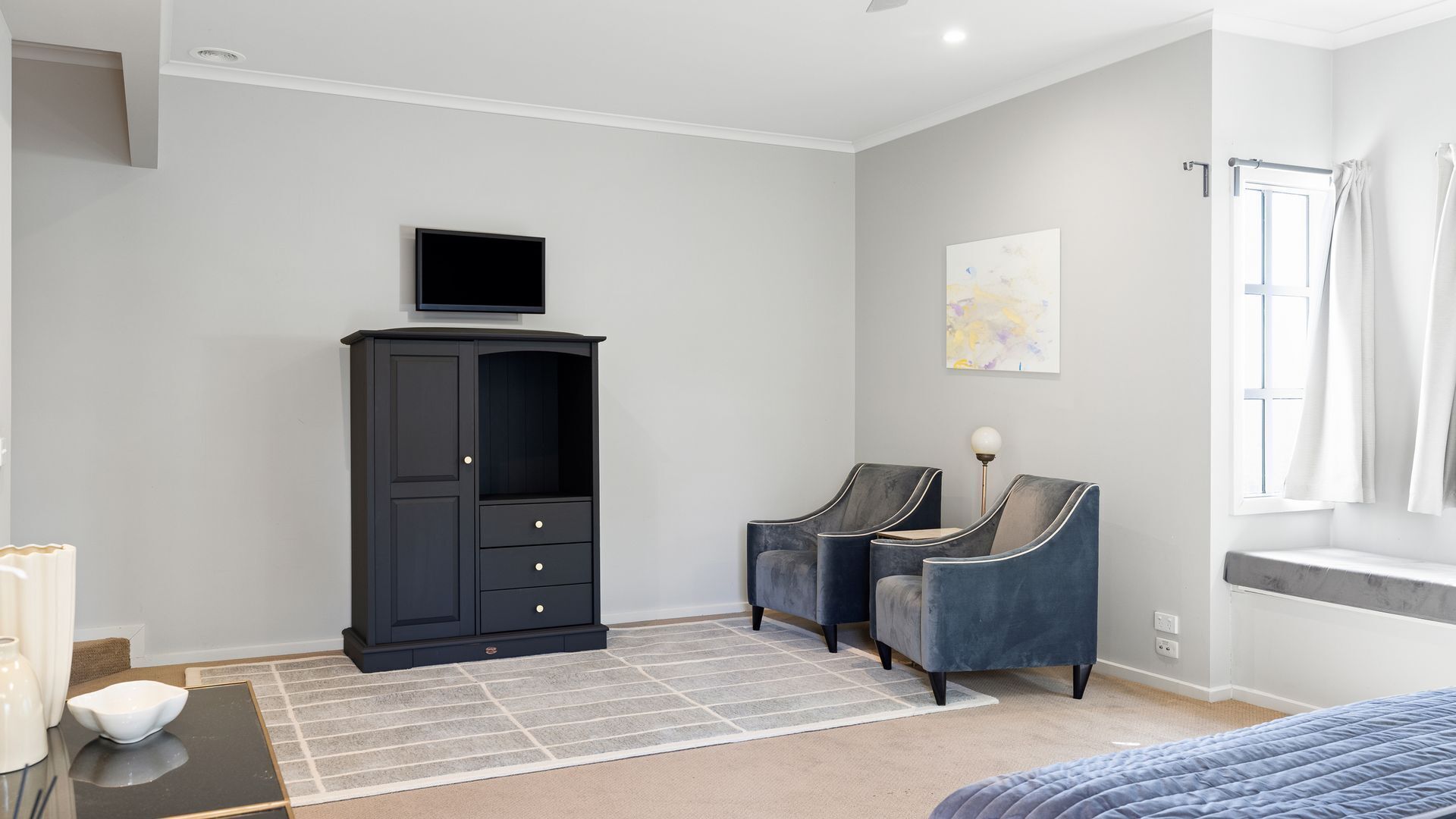 Quarto moderno com guarda-roupa escuro, duas poltronas, uma cama e uma janela com cortinas.