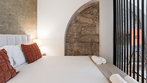 A bedroom with a stone wall, a bed with white linens, and a view through a barred window.
