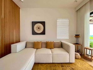 Un salon moderne avec un canapé d’angle, une œuvre d’art encadrée, une fenêtre avec des stores et une petite table d’appoint.