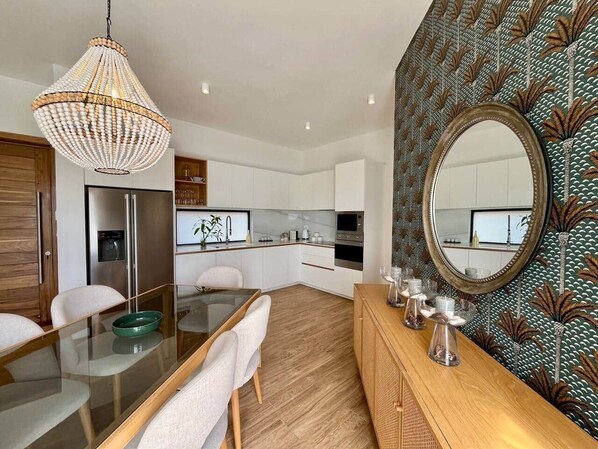 Une cuisine moderne avec des meubles blancs, un coin repas doté d’une table en verre et un miroir décoratif au mur.