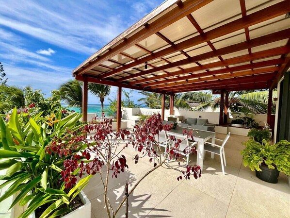 A covered outdoor seating area with a view of the ocean, surrounded by tropical plants.