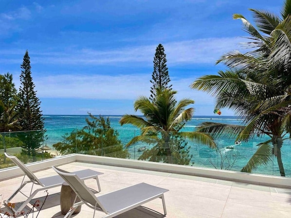 A balcony with lounge chairs overlooking a beach with palm trees and the ocean.
