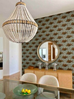 Un coin repas comprenant une table en verre, des chaises, un lustre, un miroir et un papier peint à motifs.