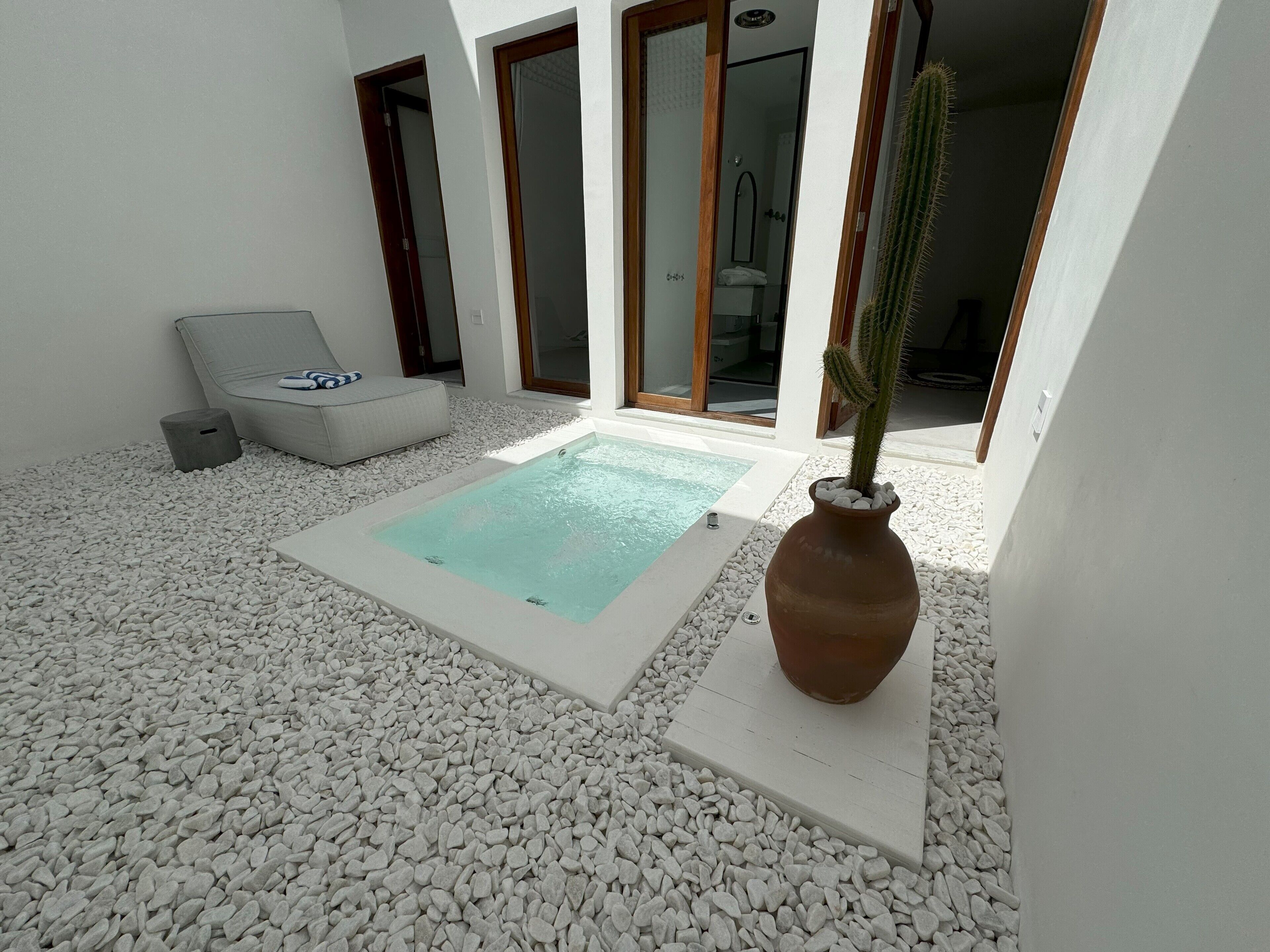 Uma área externa moderna com uma piscina retangular, pedras brancas, uma cadeira de descanso cinza e um cacto em vaso.