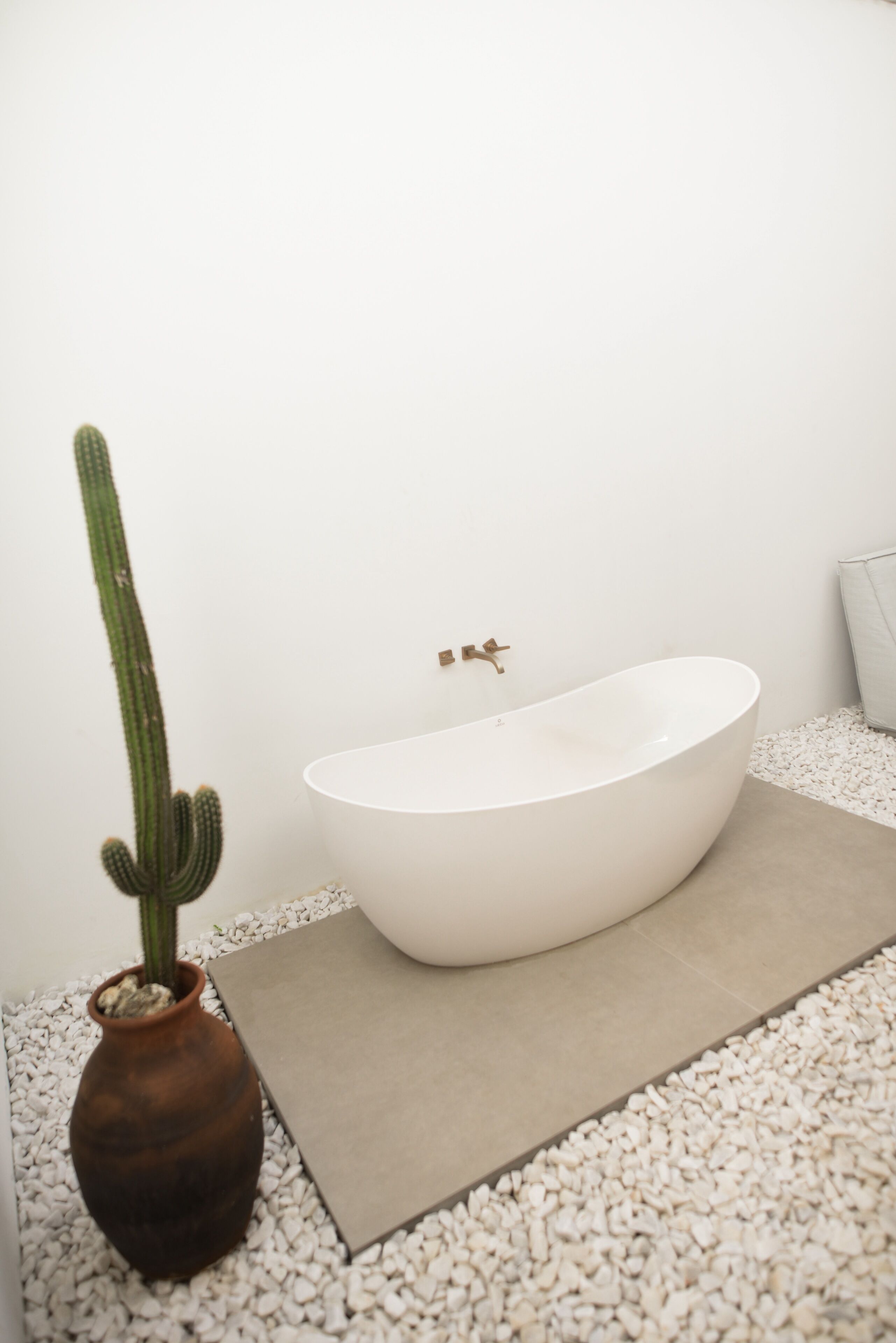A white bathtub, a cactus, and a brown vase on a white pebbled floor.