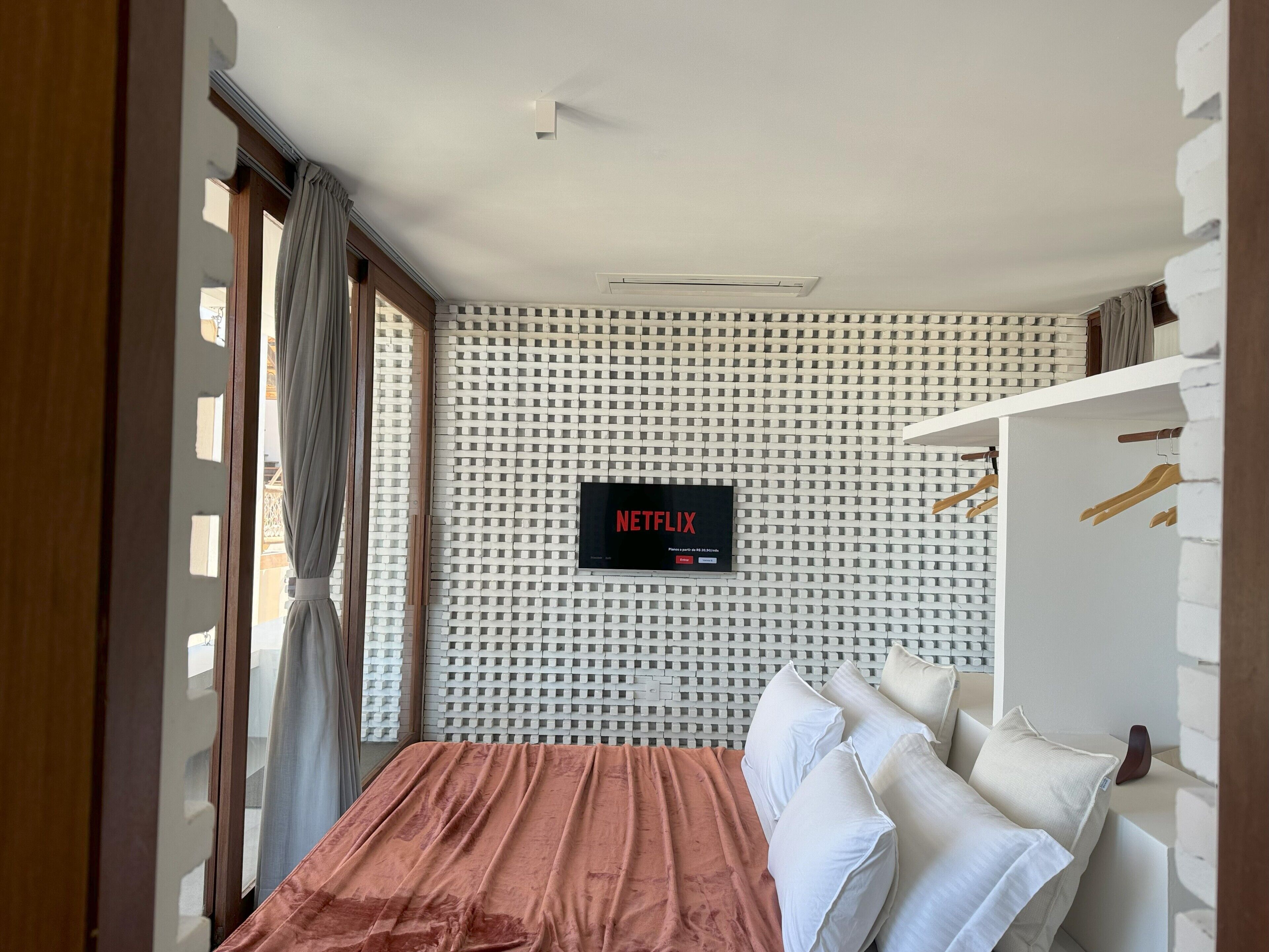 A modern bedroom with a bed, a wall-mounted TV displaying 'NETFLIX', and a white wardrobe with wooden hangers.