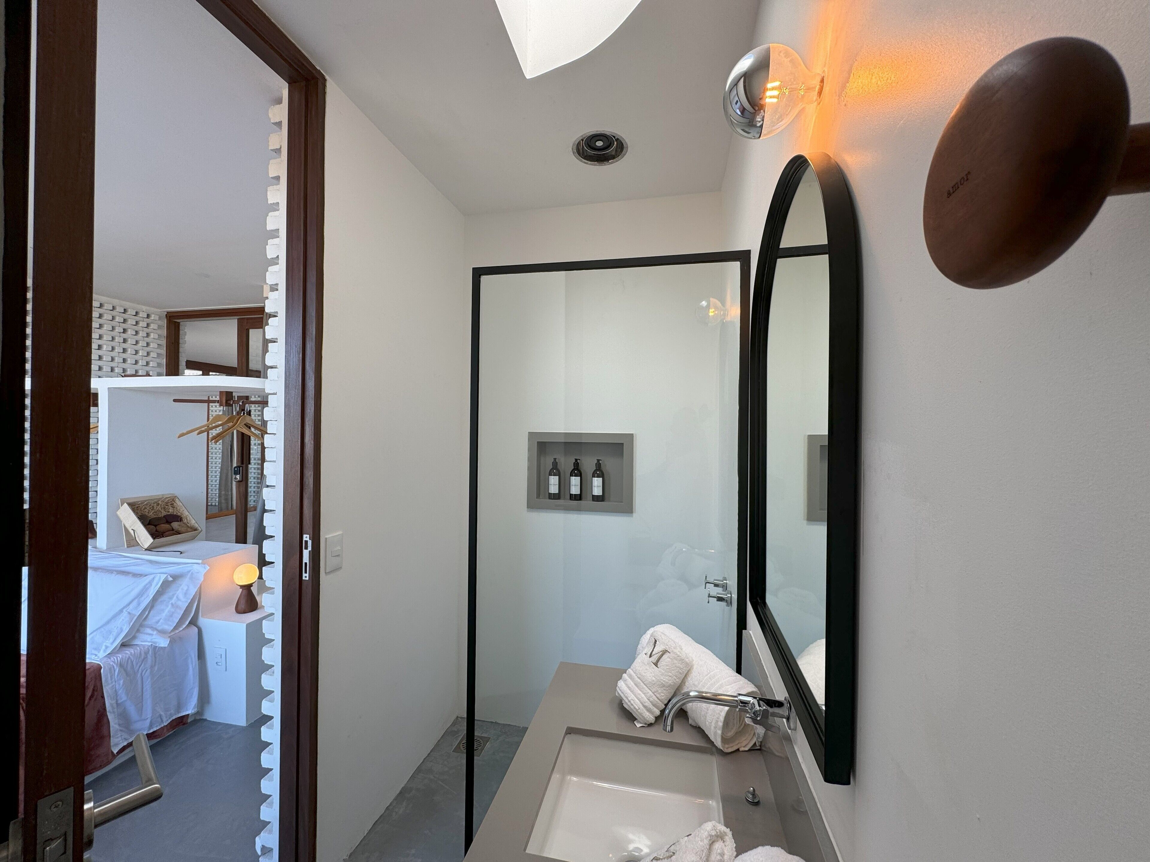 Een moderne badkamer met een grote spiegel, een wastafel en een douche. Handdoekrek met handdoeken en een klein plankje met flesjes.
