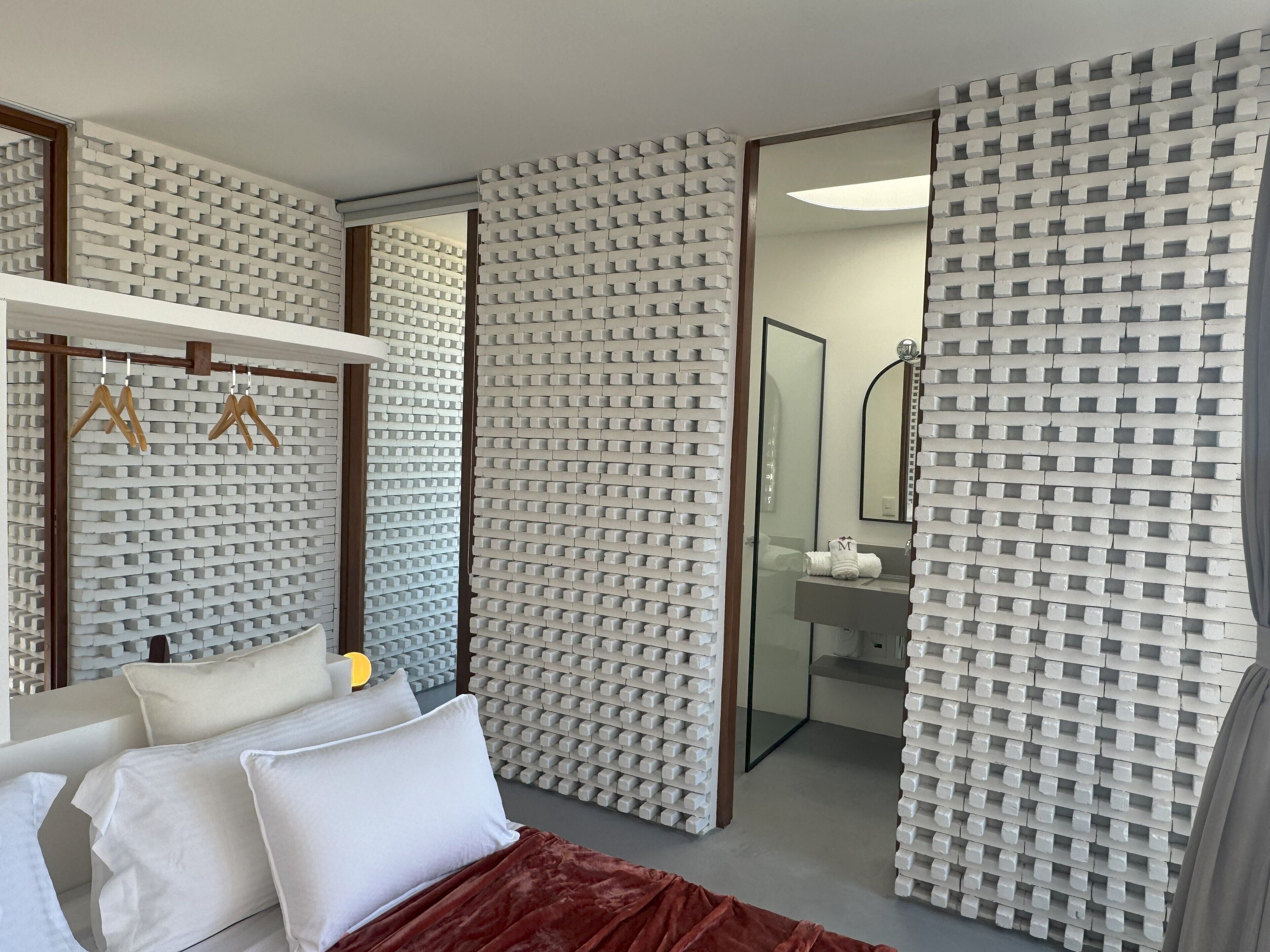 Una habitación de hotel moderna con cama, almohadas blancas, una colcha roja, un perchero de madera y un baño con espejo y lavabo.