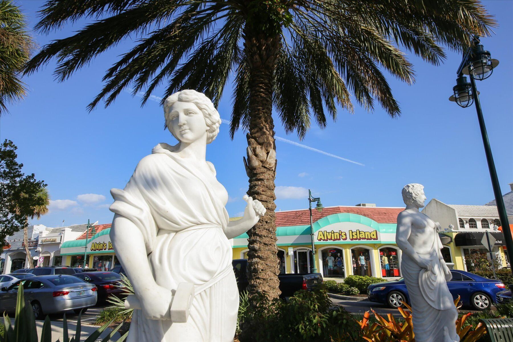Une statue représentant une femme tenant un livre, avec un palmier et un bâtiment portant l’inscription « Alvin’s Island » en arrière-plan.