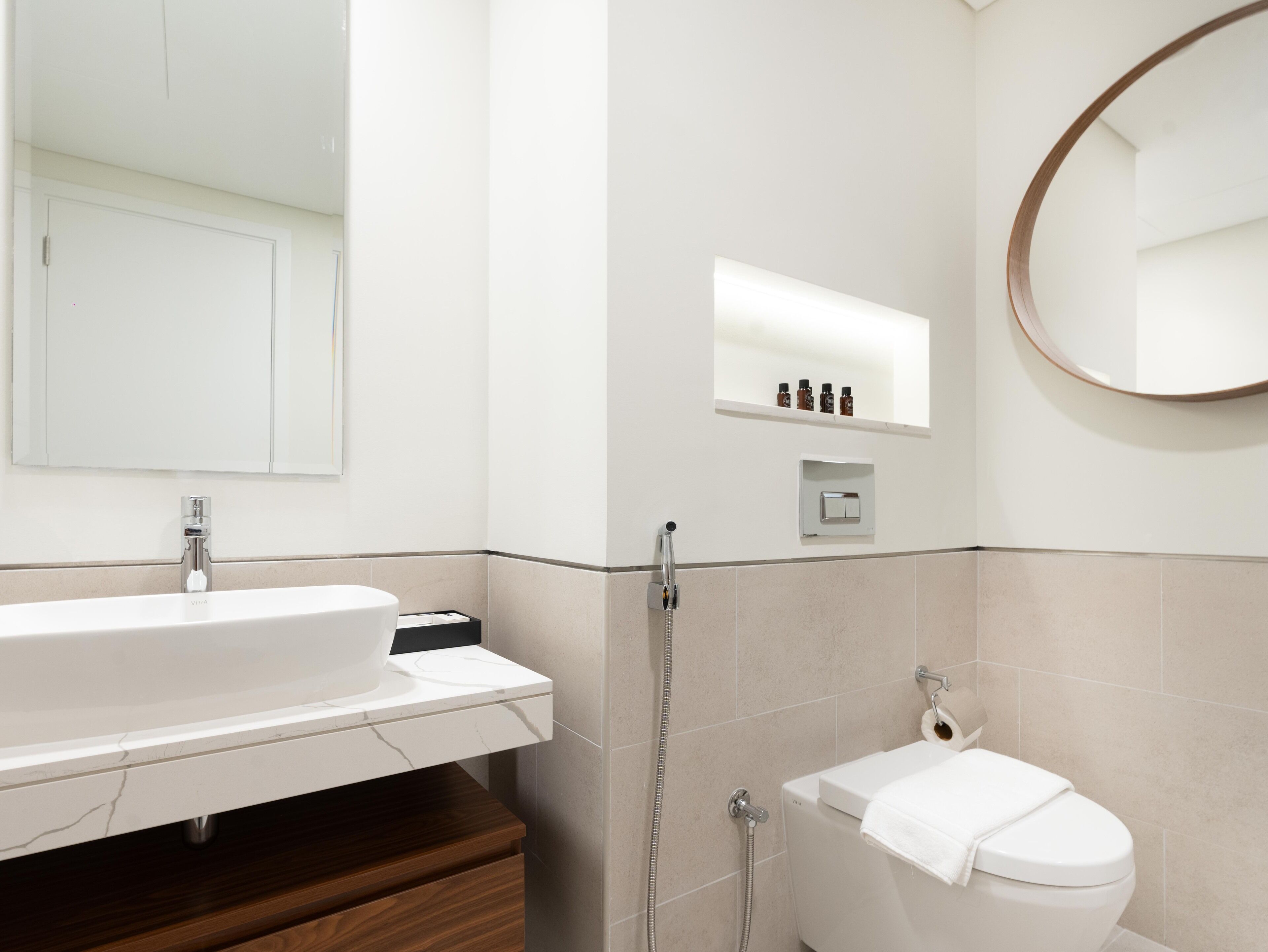 A modern bathroom with a white sink, mirror, and toilet. There is a shower area with a handheld showerhead and a towel rack.