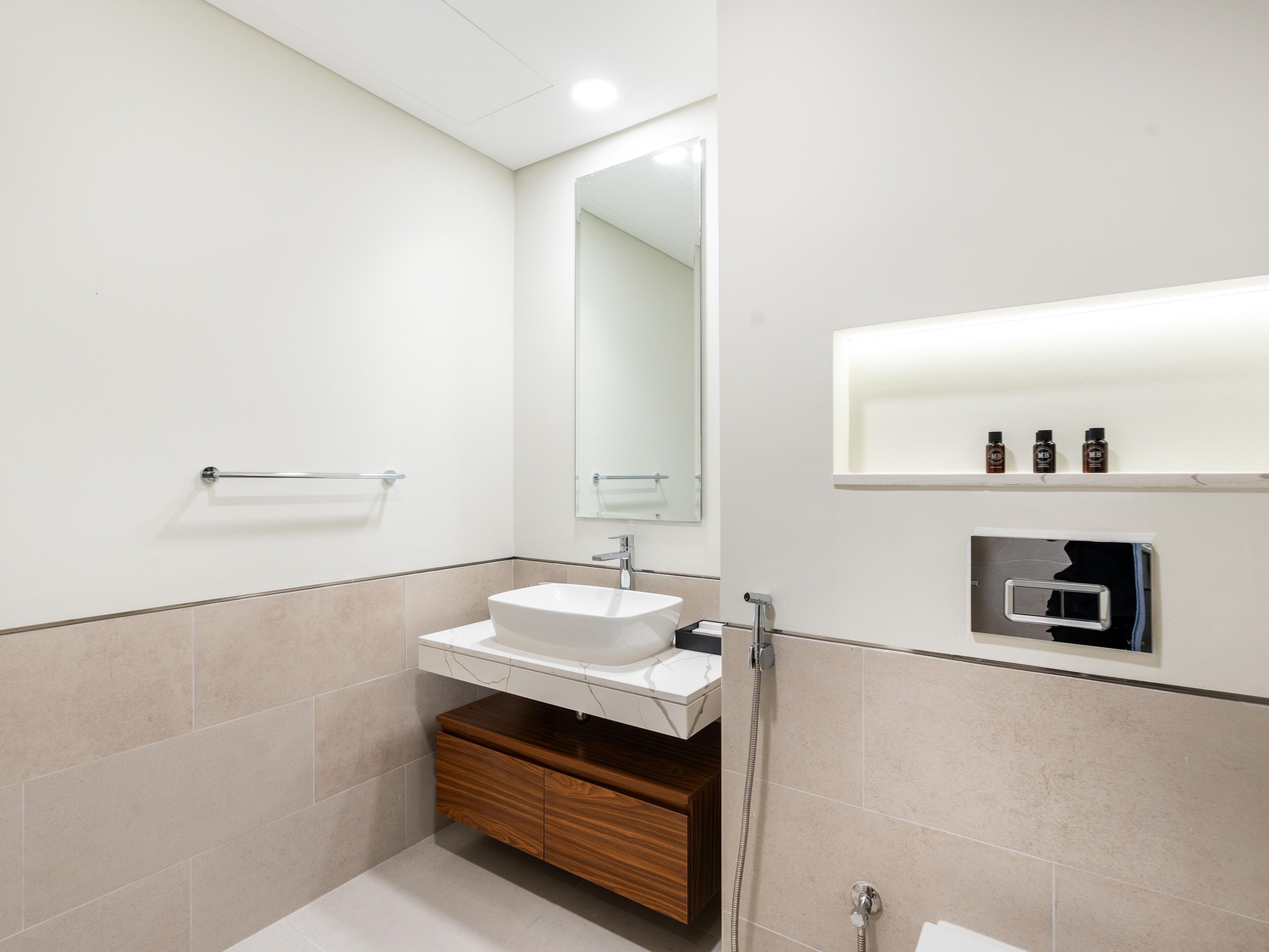 A modern bathroom with a white sink, wooden vanity, a large mirror, and a shelf with toiletries.