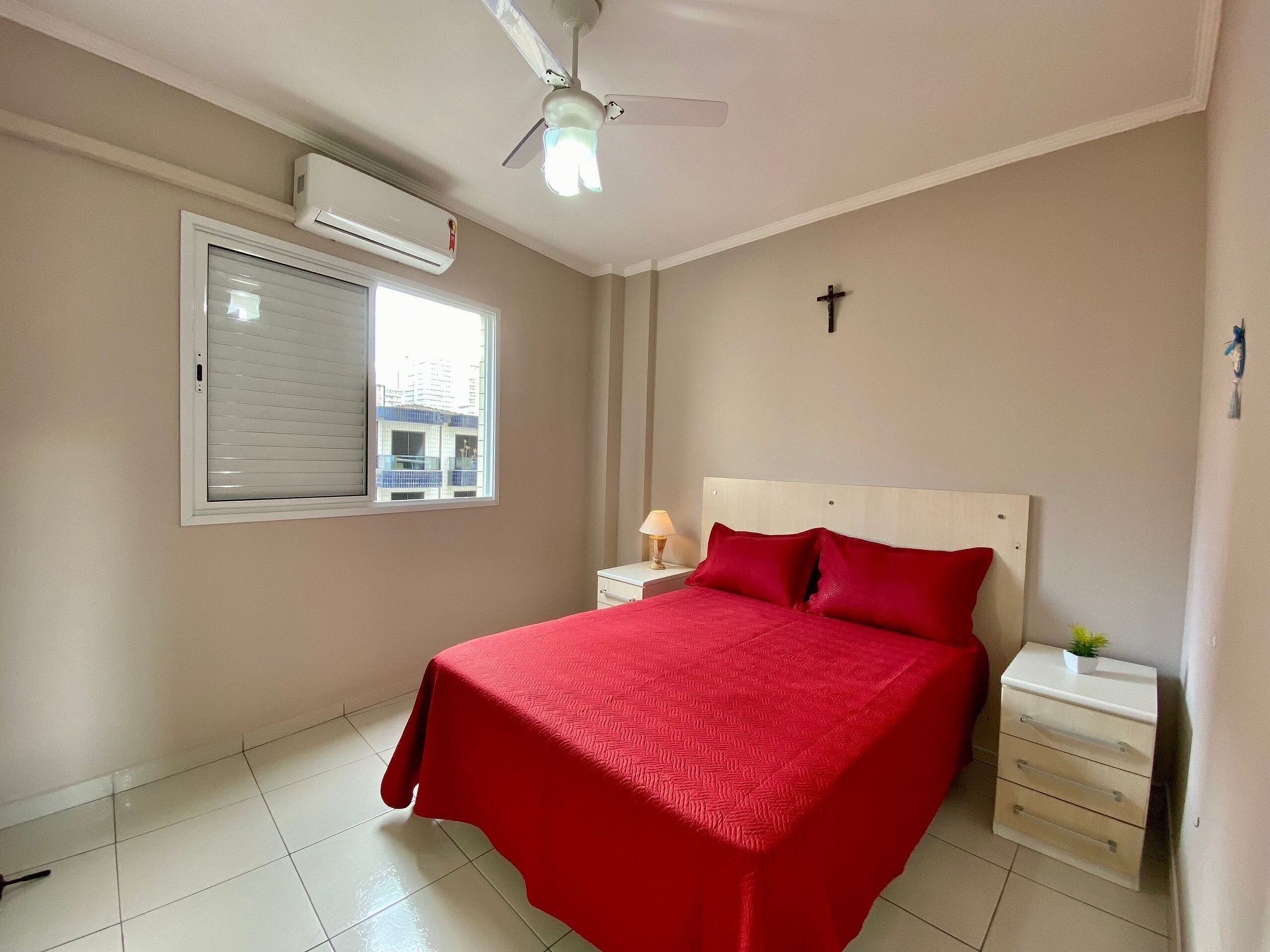 A bedroom with a bed, a nightstand, a lamp, a ceiling fan, an air conditioning unit, and a window.