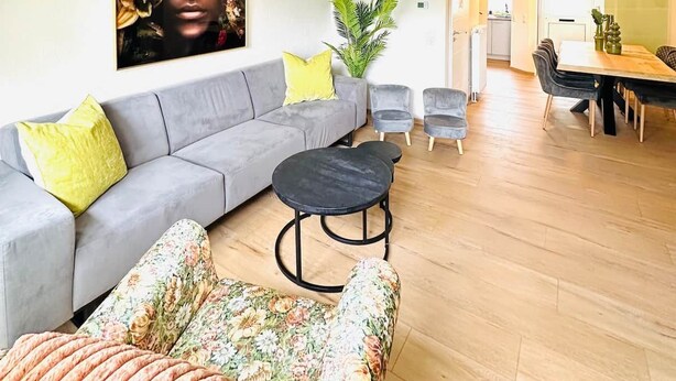 Een moderne woonkamer met een grijze bank, een fauteuil met bloemenprint en een ronde zwarte tafel. Op de achtergrond is een eethoek met een houten tafel en stoelen zichtbaar.