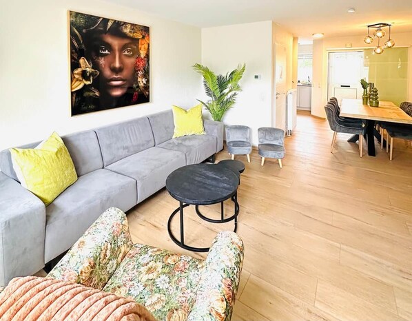 A modern living room with a grey sofa, floral armchair, and a round black table. A dining area with a wooden table and chairs is visible in the background.
