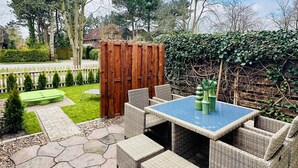 Een terras buiten met een tafel en stoelen, omringd door een houten schutting en begroeiing.