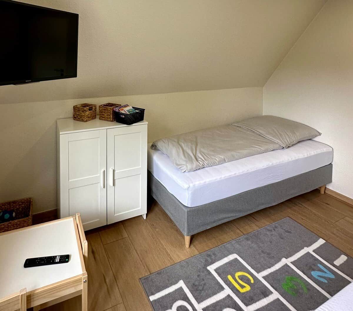 Ein kleines Zimmer mit einem Einzelbett, einem weißen Kleiderschrank, einem an der Wand befestigten Fernseher und einem kleinen Tisch mit einer Fernbedienung.
