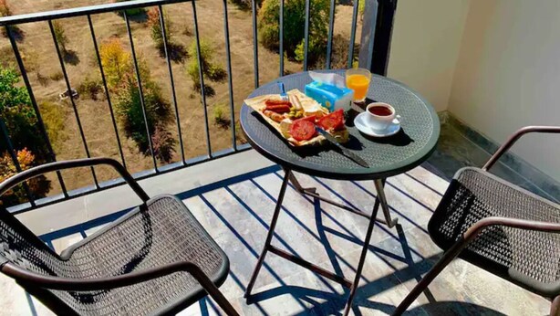 Ein Balkon mit zwei Stühlen, einem Tisch mit Frühstücksgebäck und Blick auf Bäume und eine Rasenfläche.