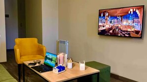 Een hotel lobby met een gele fauteuil, een houten tafel met een laptop, een televisie aan de muur en een groene poef.