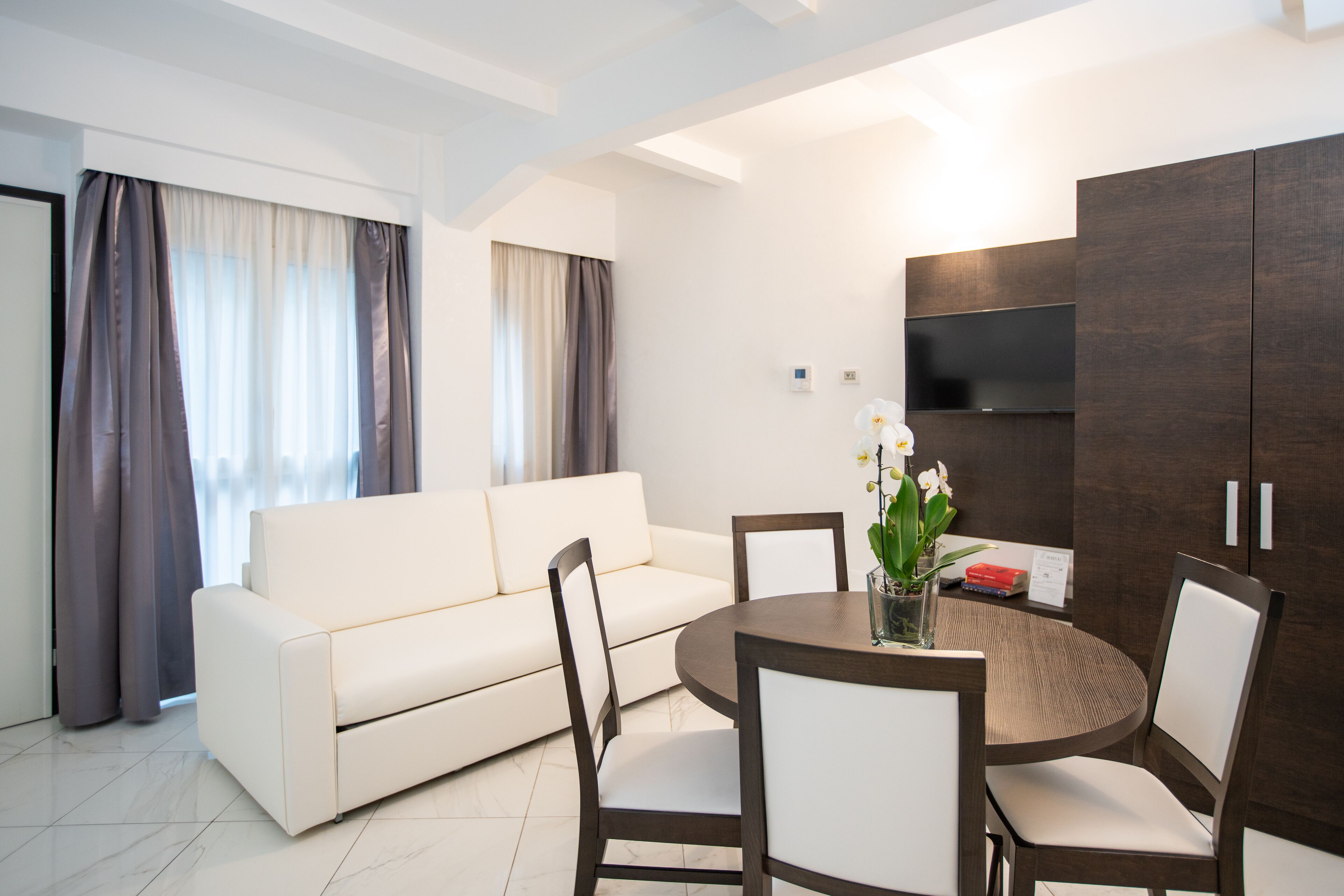 A modern living room with a white sofa, a round wooden dining table, and matching chairs. There is a dark wooden cabinet with a flat-screen TV mounted above it.