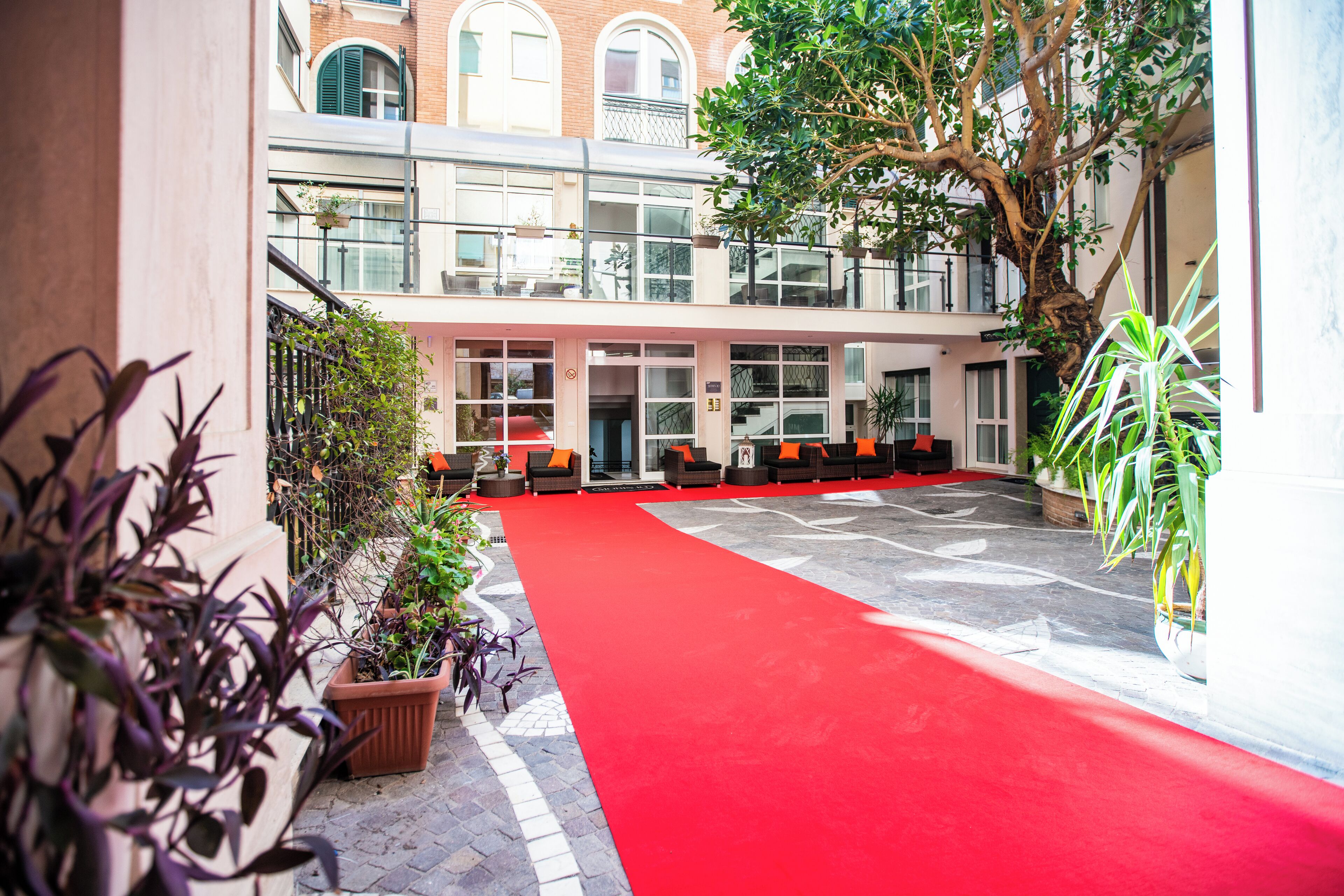 A red carpeted area with seating and potted plants, a staircase, and a building with large windows and balconies.