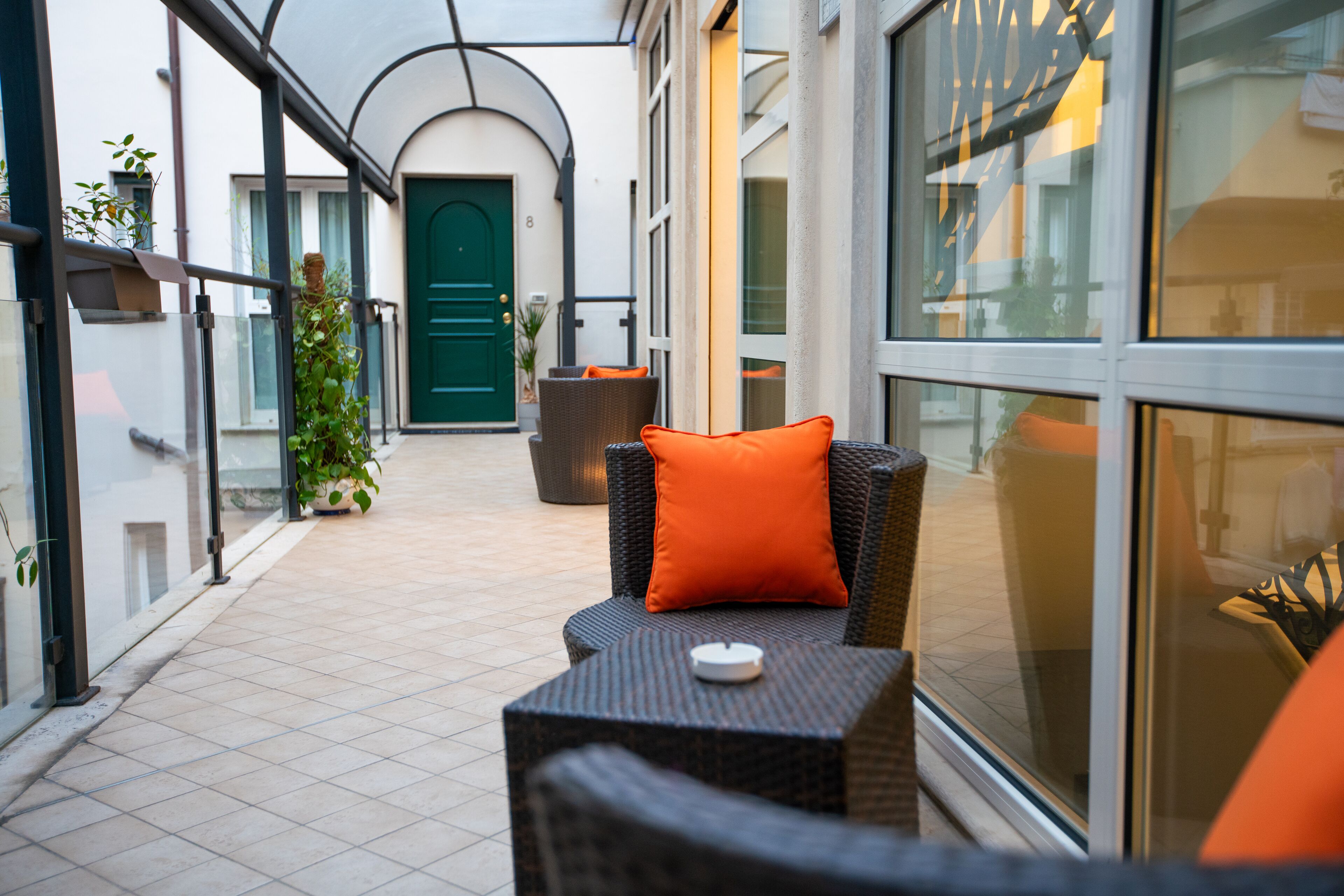 An outdoor seating area with wicker furniture and orange cushions, a green door in the background, and potted plants along the walkway.