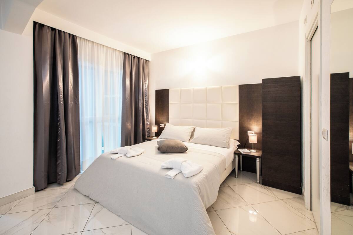 Ein modernes Hotelzimmer mit einem großen Bett, dunklen Vorhängen, einem Nachttisch mit Lampe und einer Tür rechts.