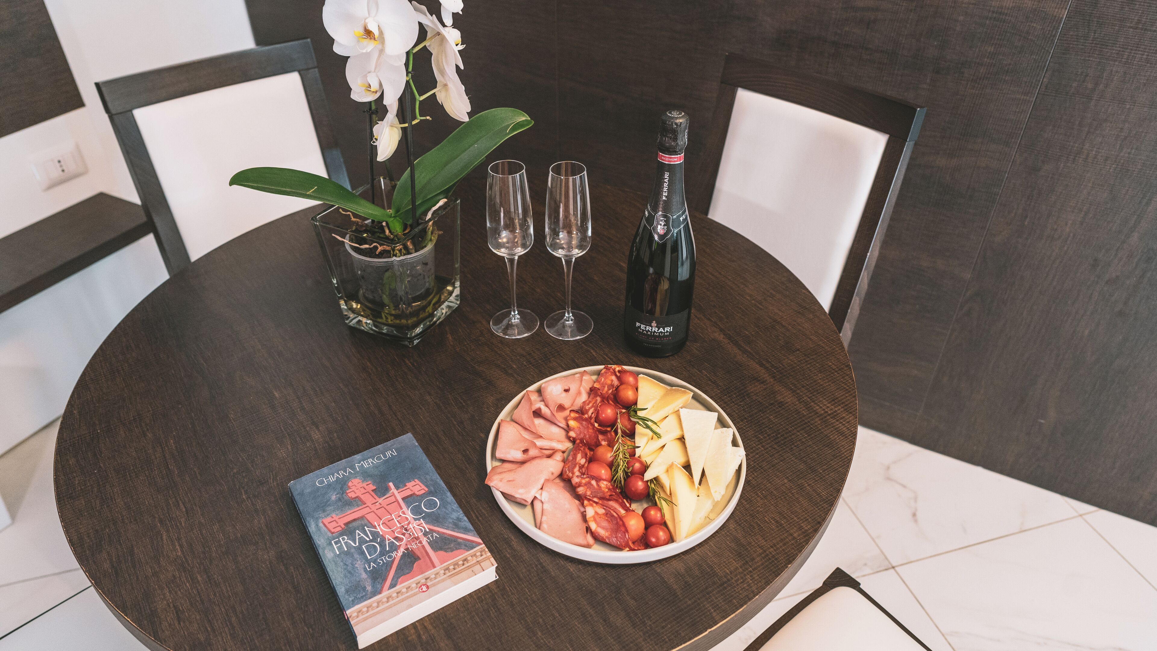 A round table with a book, a plate of food, two glasses, a bottle, and an orchid.