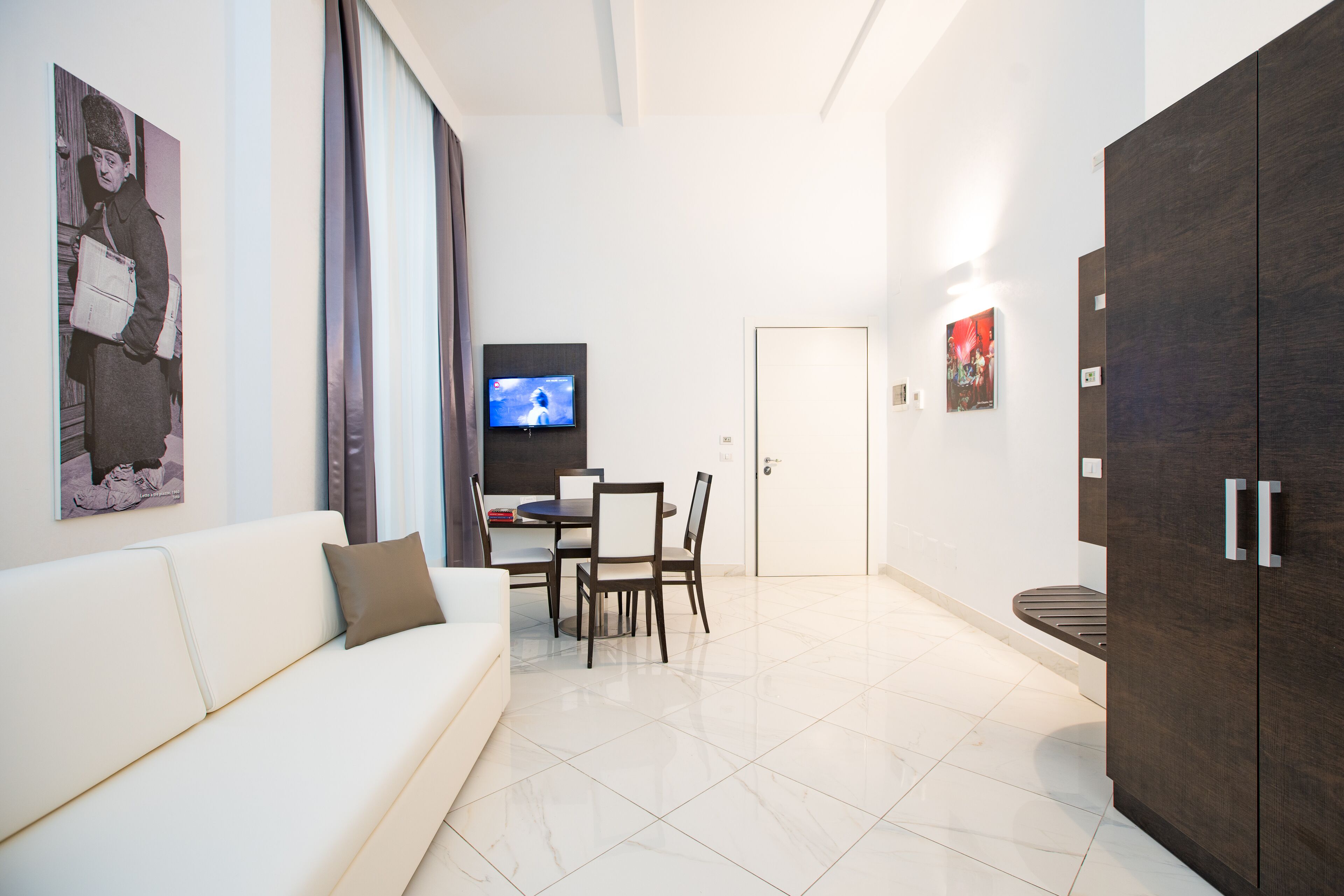 A modern living room with a white sofa, a dining table with chairs, a TV on the wall, and a dark wooden wardrobe.