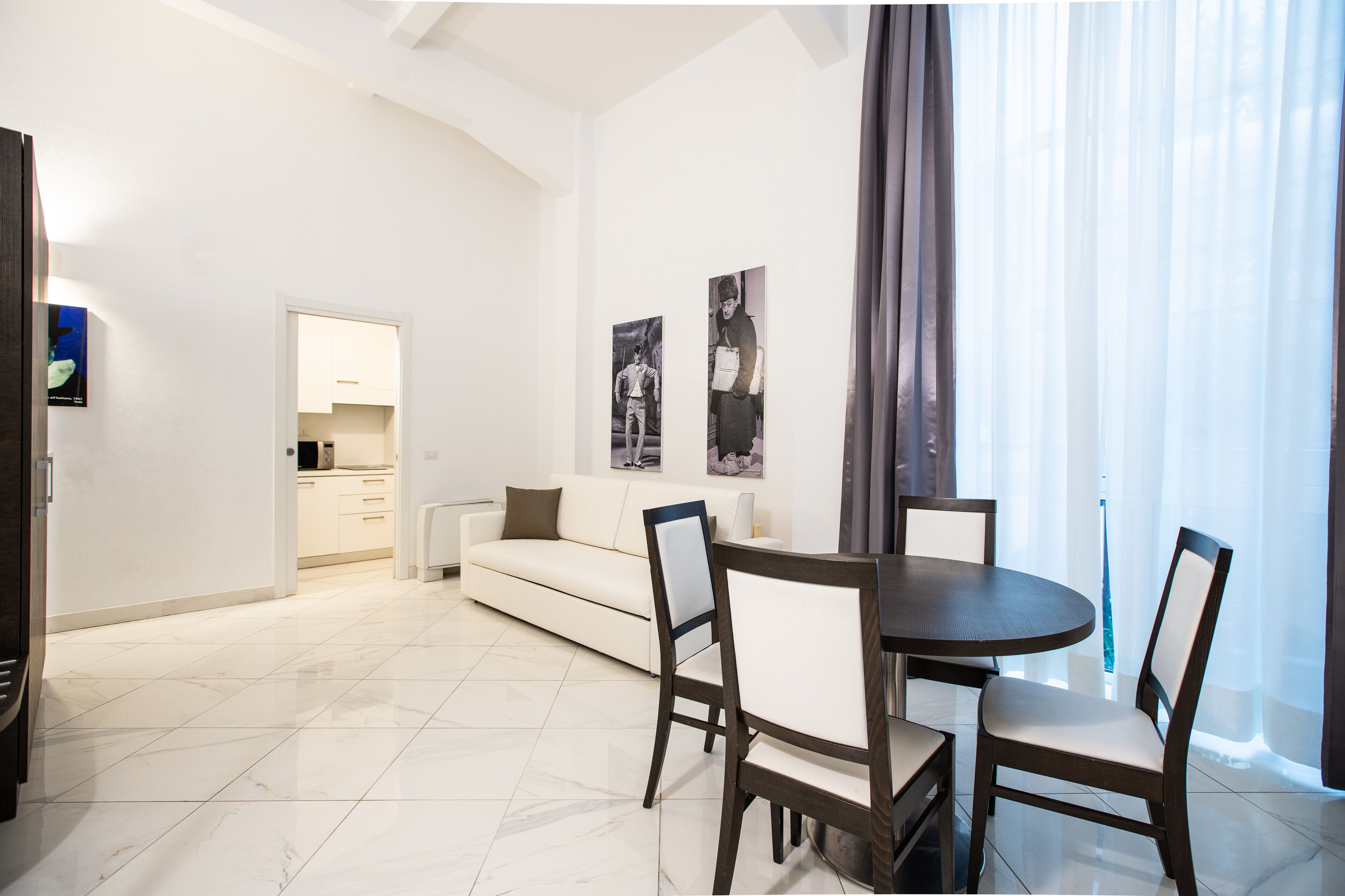 A modern living room with a dining area, white walls, and a large window with sheer curtains.