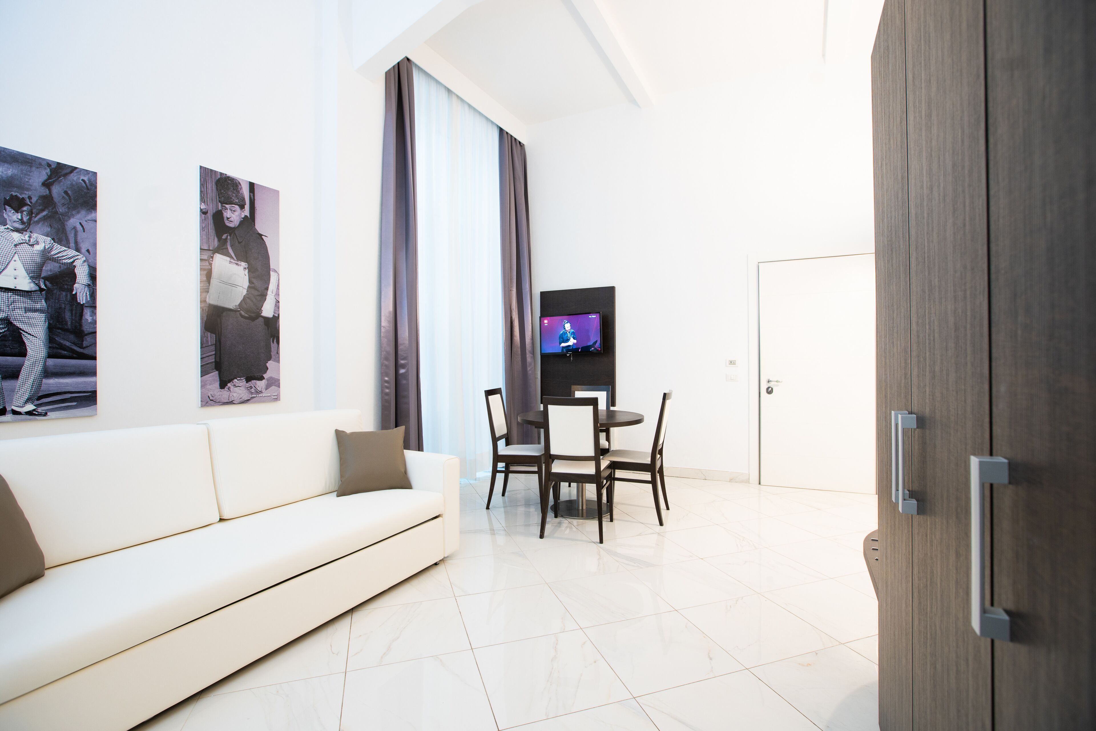 A modern living room with a white sofa, a dining table with chairs, and a flat-screen TV mounted on the wall.