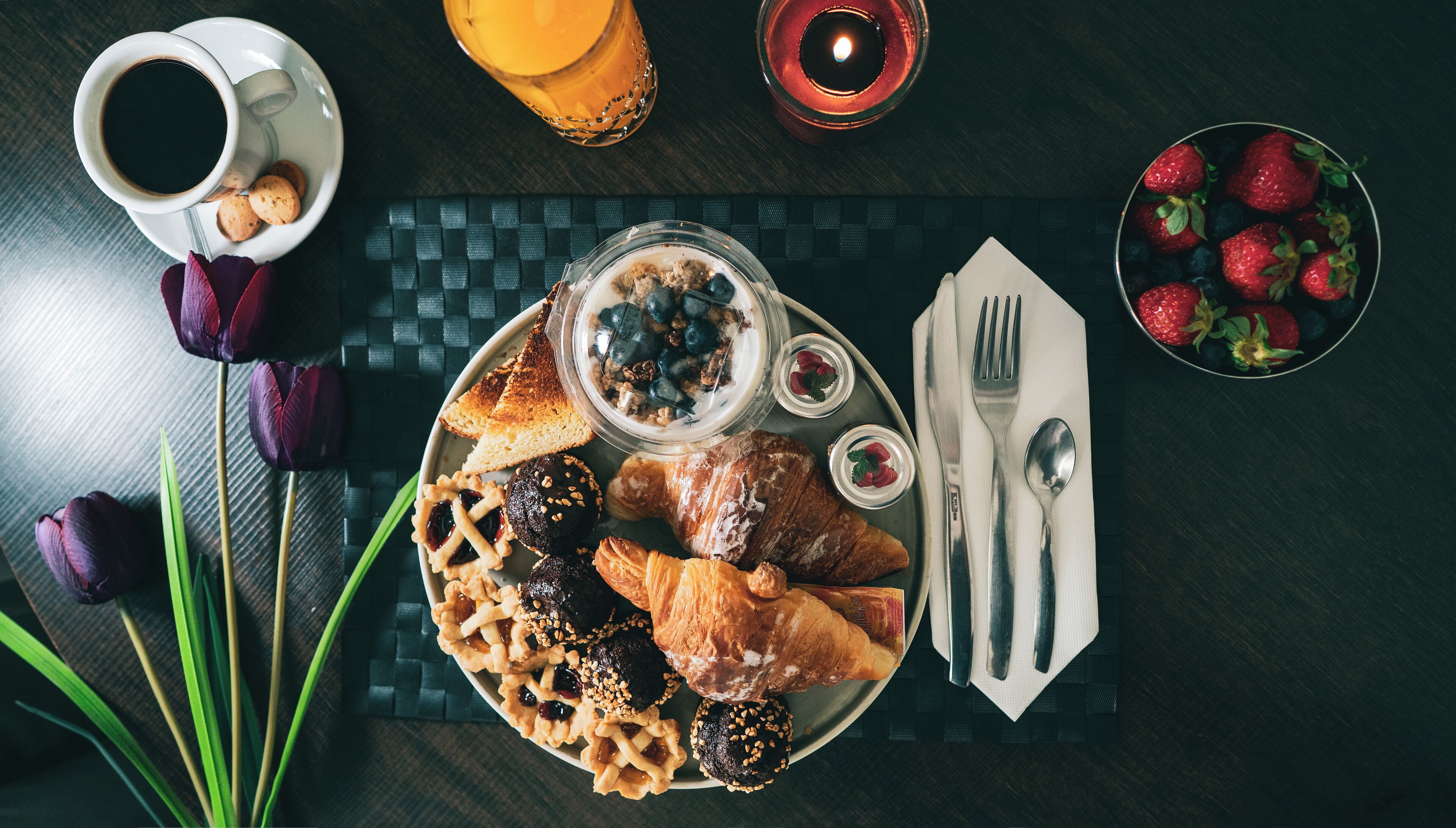 A breakfast setting with croissants, pastries, a bowl of fruit, a cup of coffee, and a candle on a dark wooden table.
