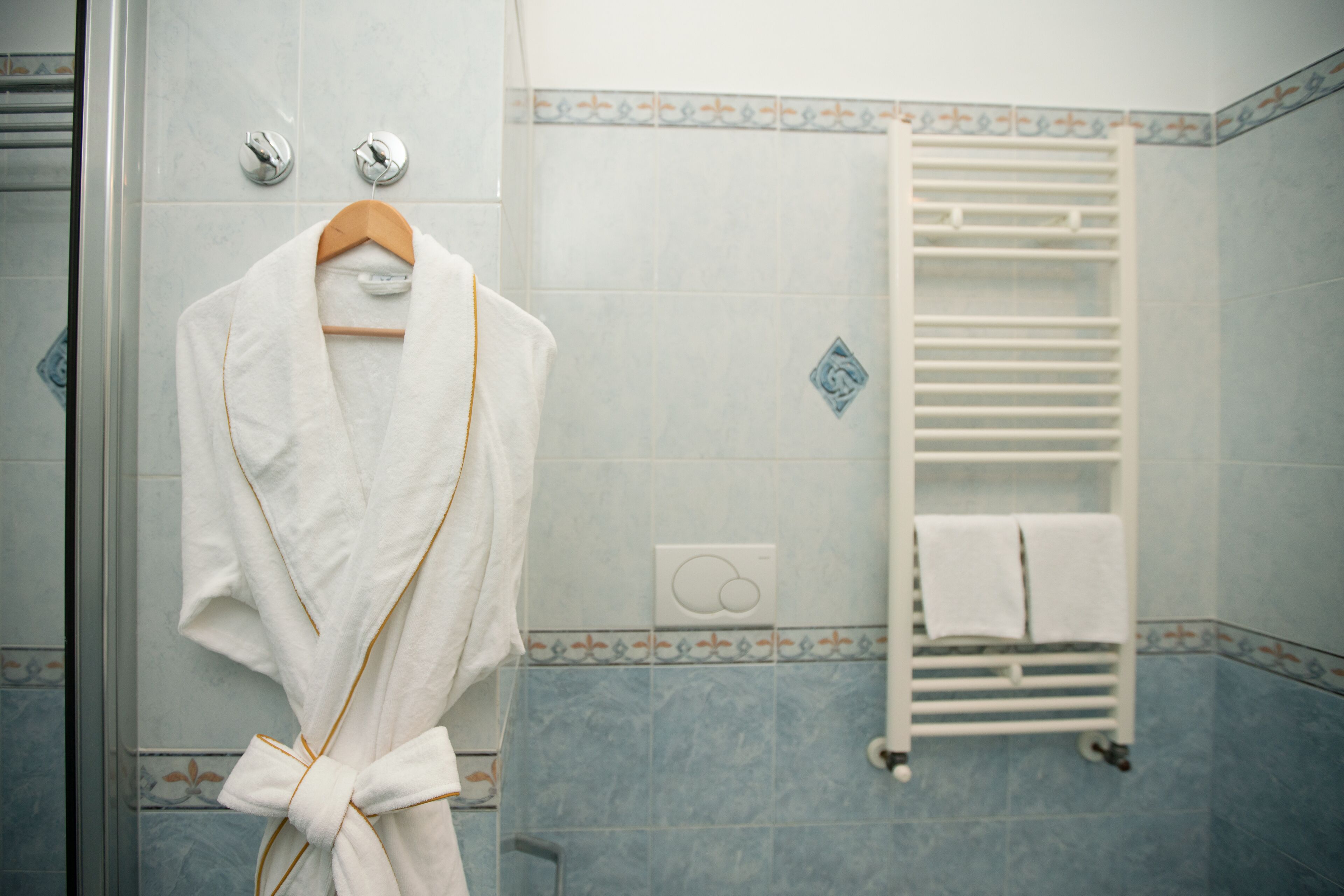A bathroom with a white bathrobe hanging on a wooden hanger, a white towel rack with two towels, and a shower with a glass door.