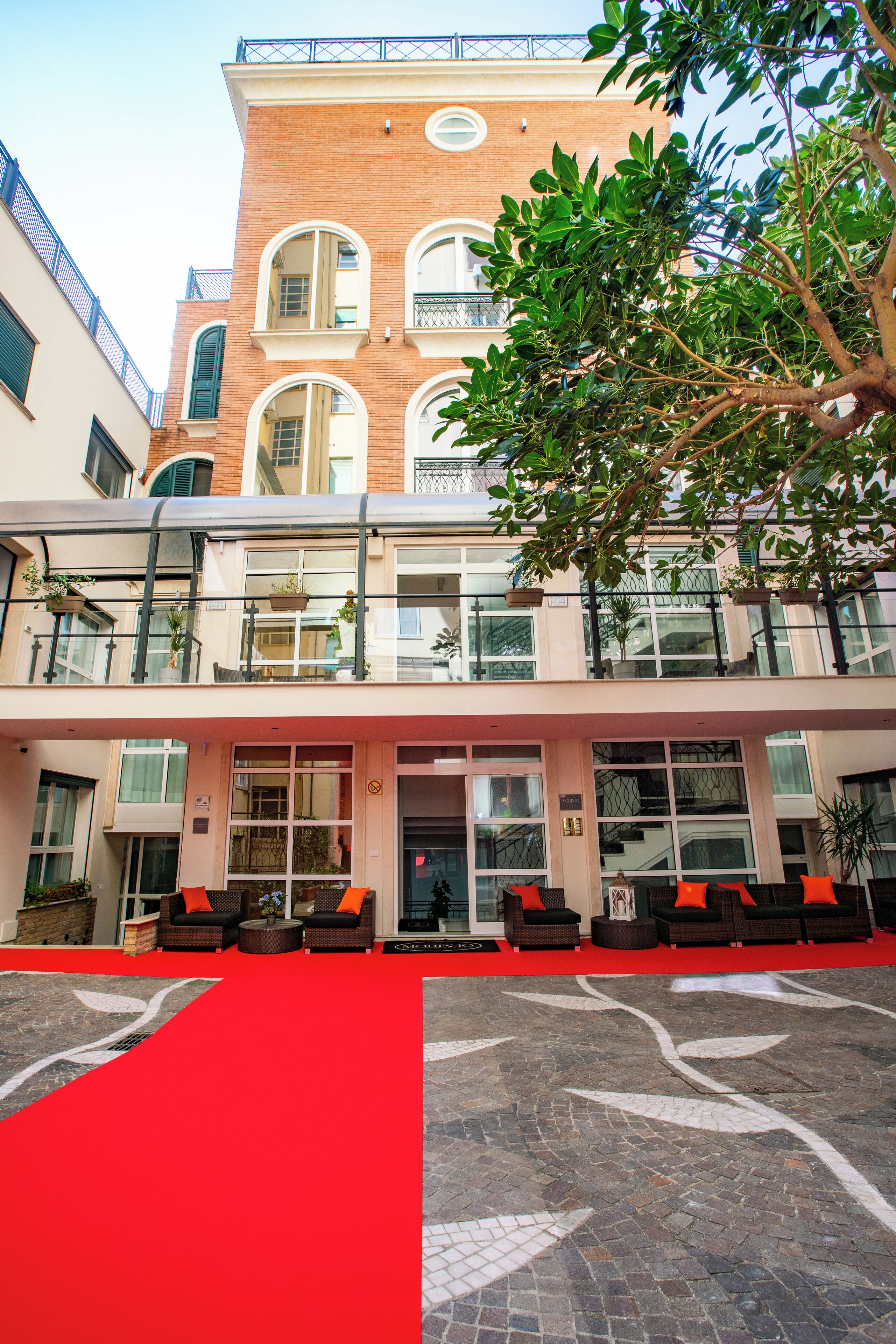 Um hotel com entrada de tapete vermelho, área externa com sofás e fachada de tijolos.
