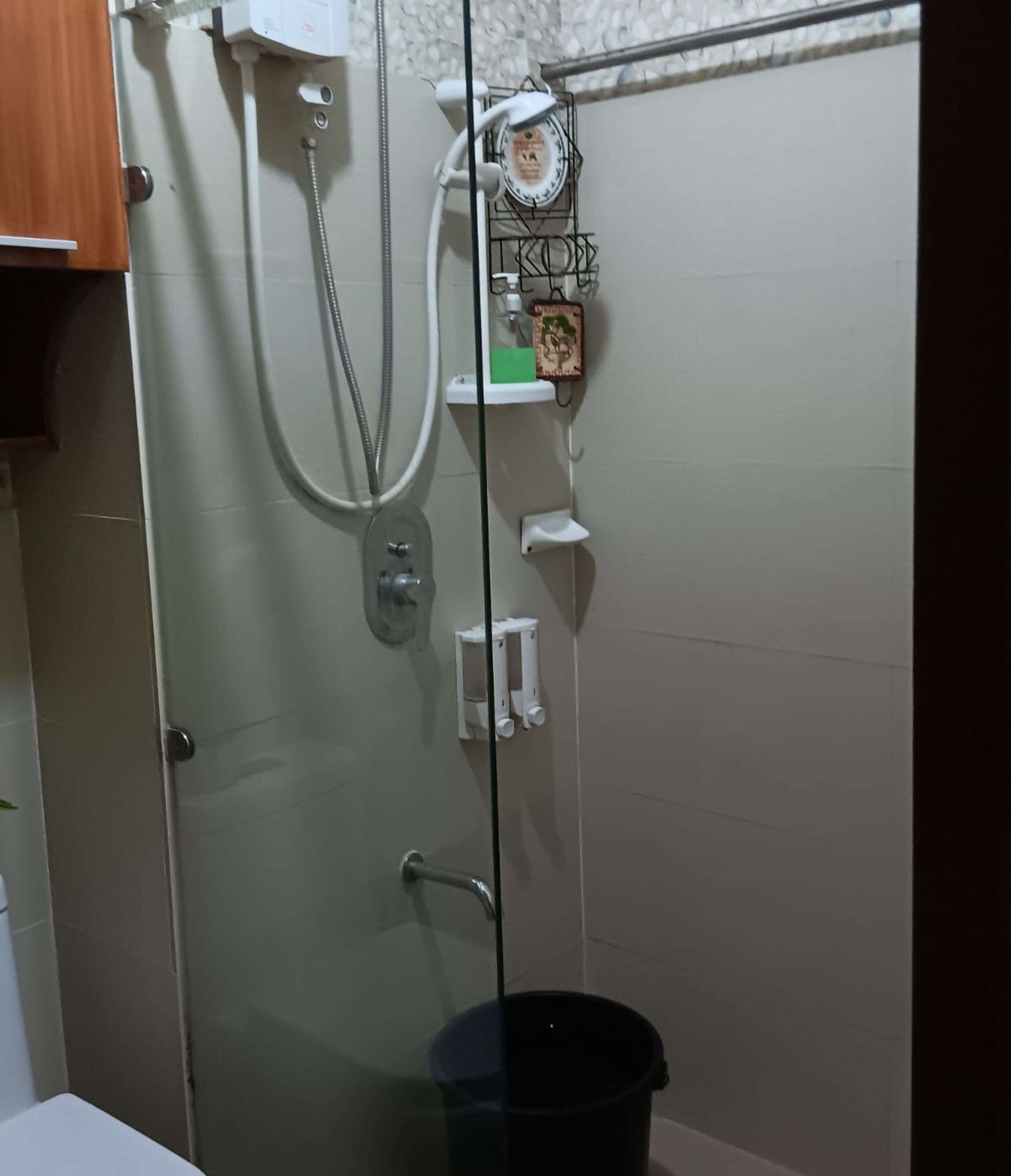 A shower area with a glass door, a white tiled wall, a showerhead, a soap dispenser, and a black bucket.