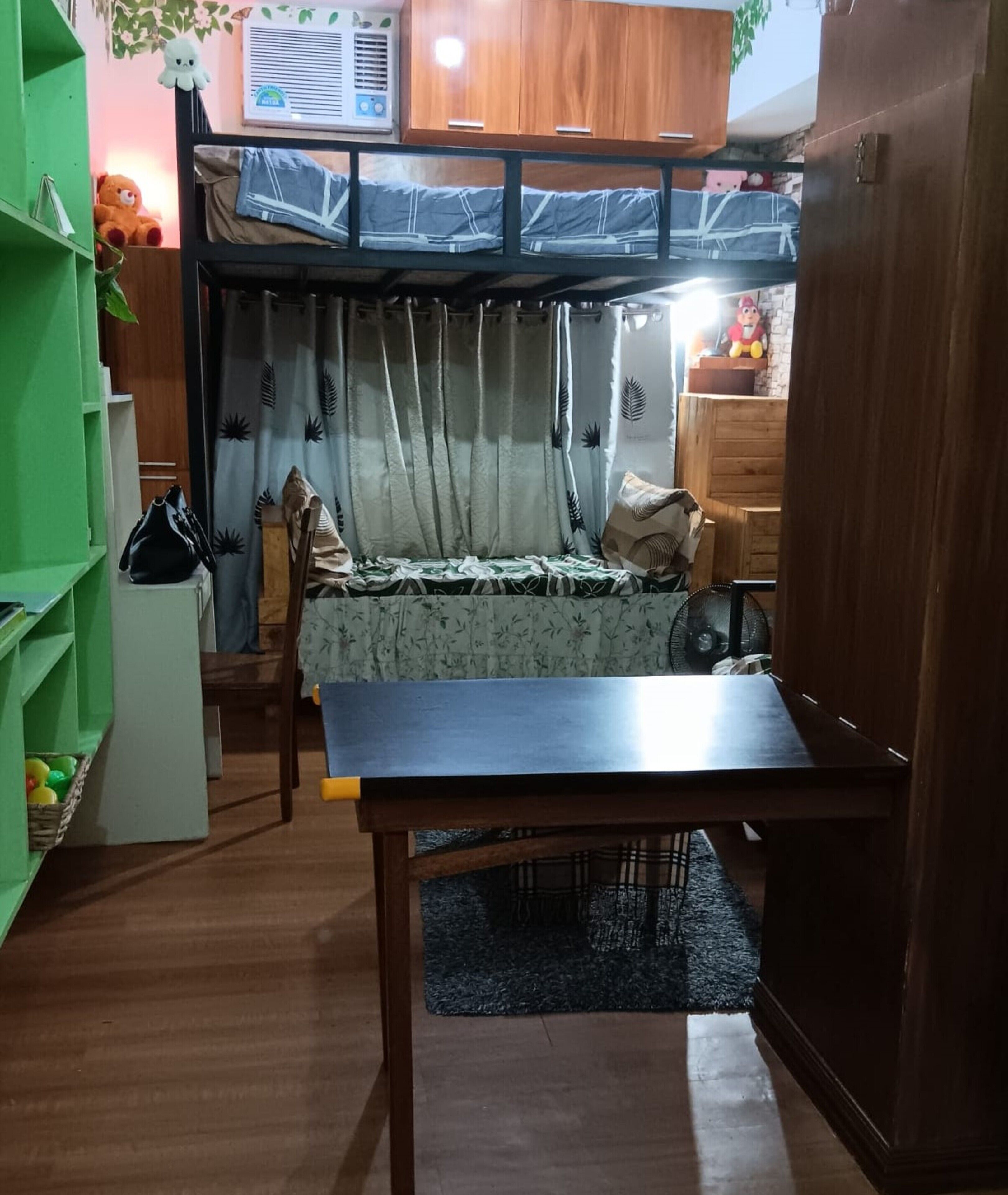 Een kleine kamer met een stapelbed, een klein tafeltje, een stoel, een groen wandrek en een ventilator.