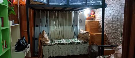 A dormitory room with bunk beds, a desk, a chair, a fan, and a small table.