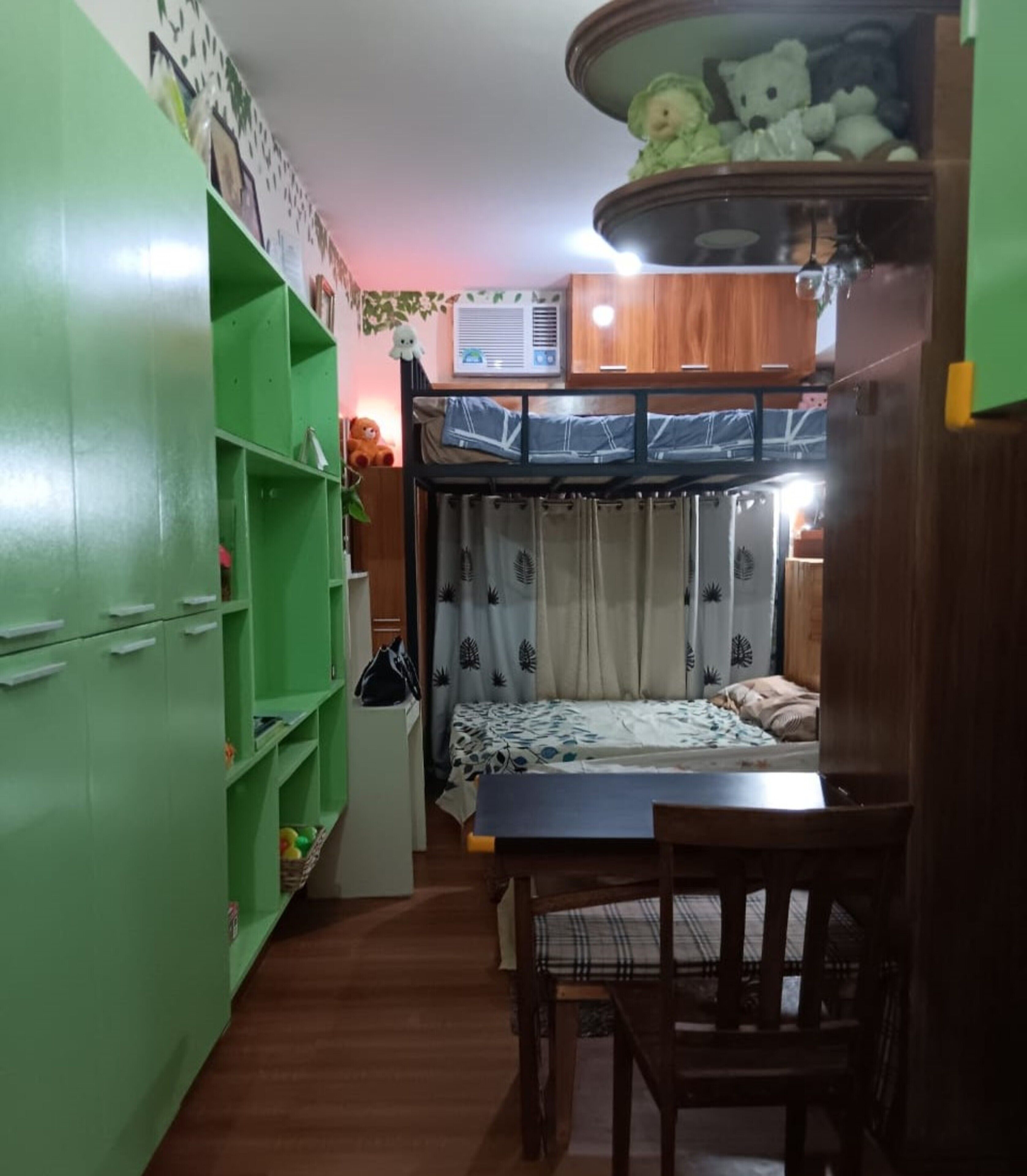 Une petite pièce comprenant un lit superposé, un petit bureau avec une chaise, une armoire verte et une étagère.