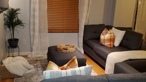 A modern living room with a dark grey sofa, a small side table, a potted plant, and a large window with blinds. - Cozy 2 Bedroom with King Bed, 2 Full Bathrooms Conveniently located in the heart of Muskegon!!! (Muskegon)