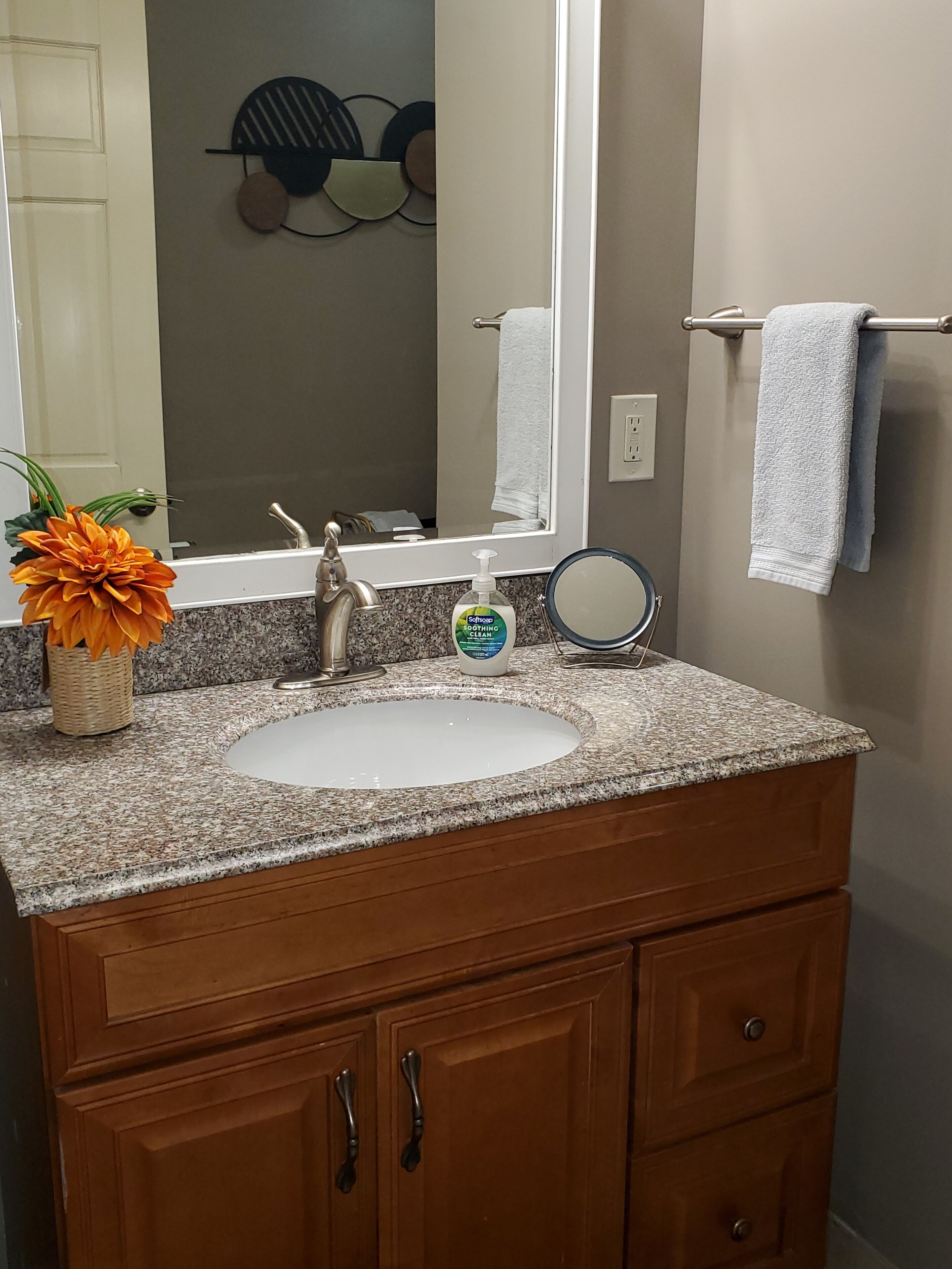 Une salle de bain dotée d’un comptoir en granit, d’un lavabo blanc, d’armoires en bois, d’un miroir, d’un porte-serviettes et d’un arrangement floral décoratif aux couleurs orangées.