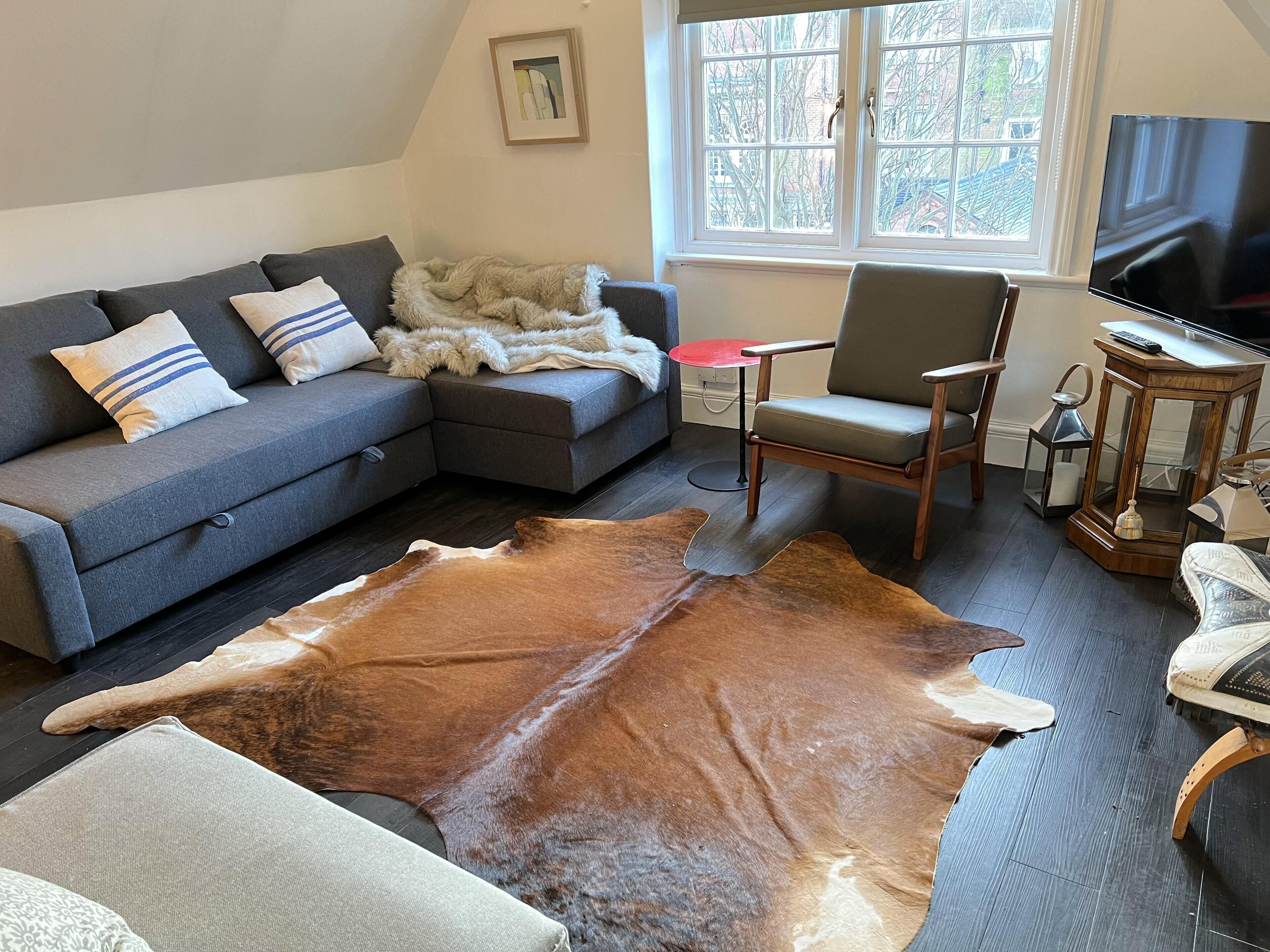 A living room with a grey sectional sofa, a wooden coffee table, a leather rug, a grey armchair, and a TV mounted on the wall.