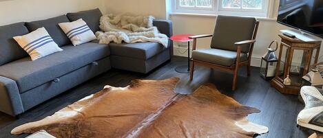 A living room with a grey sectional sofa, a wooden coffee table, a leather rug, a grey armchair, and a TV mounted on the wall.