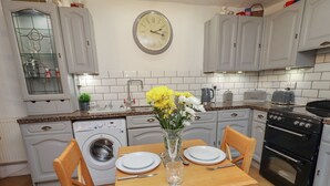 A kitchen with white cabinets, a clock, a washing machine, and a dining table set with plates and a vase of flowers.