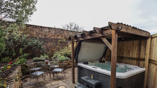 A hot tub under a wooden pergola in a garden with stone flooring, surrounded by potted plants and a brick wall.
