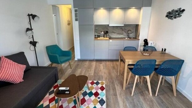 A compact living space with a sofa, a coffee table, a patterned rug, a dining area with a table and chairs, and a kitchenette with cabinets and a countertop.