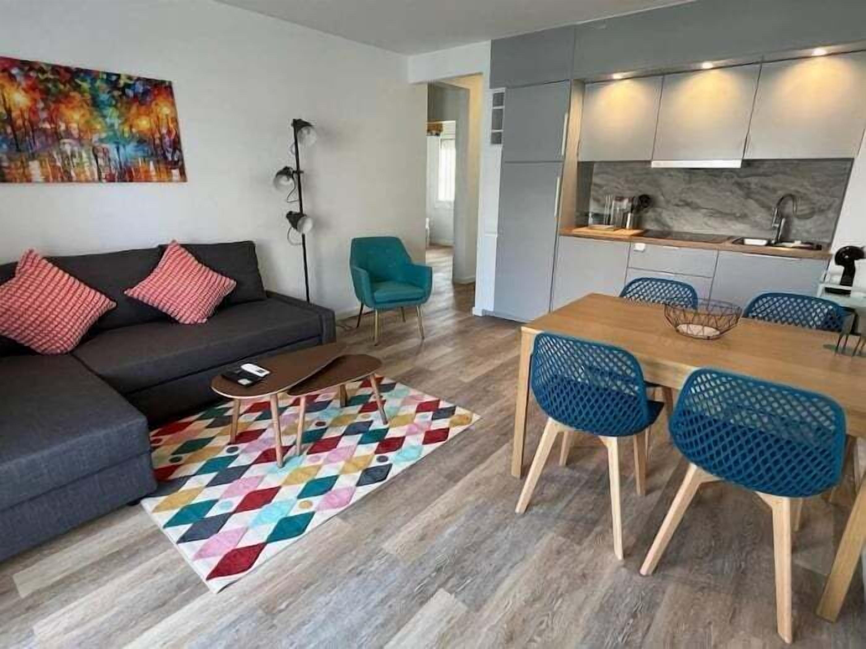A modern living room with a sofa, a coffee table, and a colorful rug. There is a dining area with a wooden table and blue chairs.