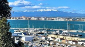 A marina with boats docked, a coastal cityscape with buildings, and snow-capped mountains in the background.